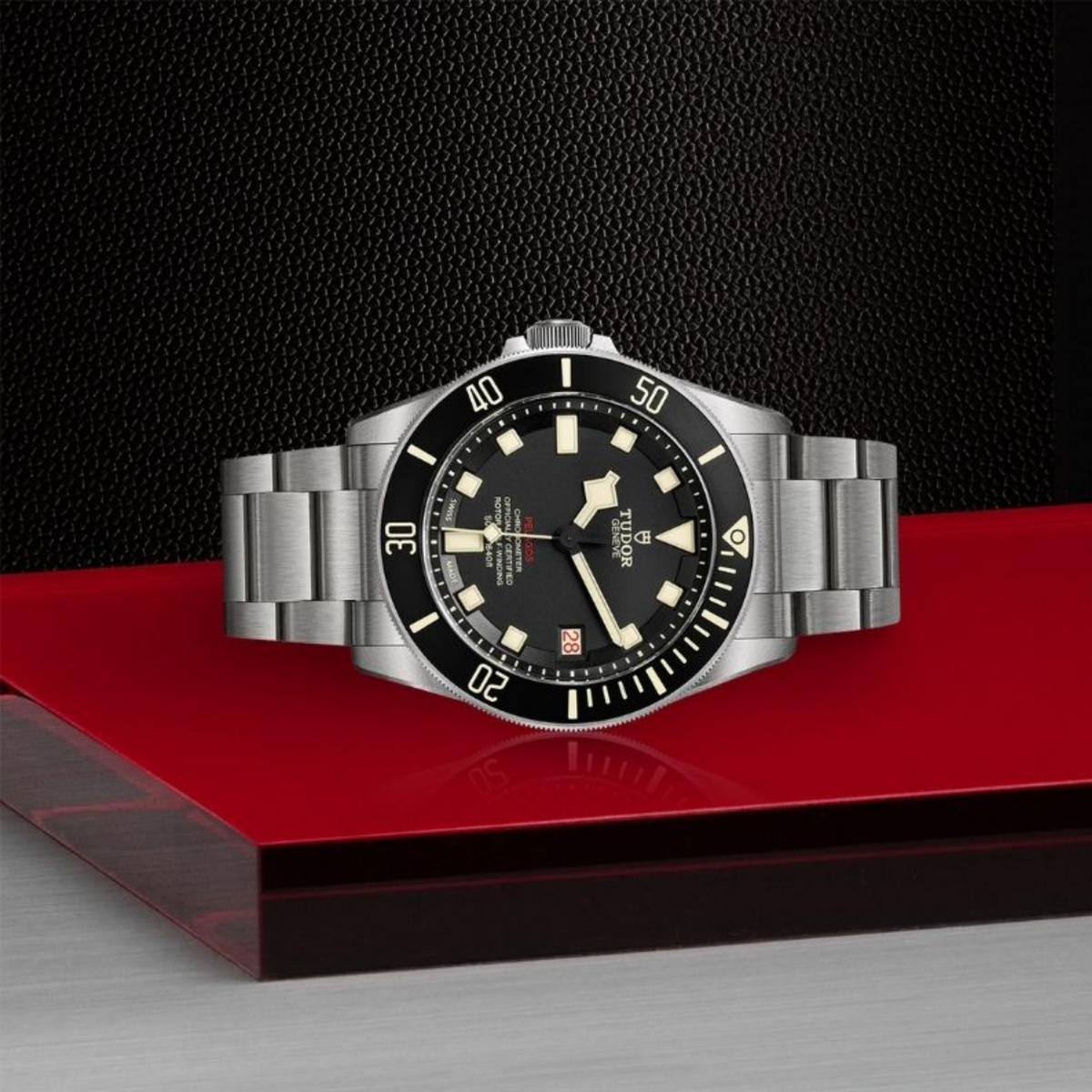 Tudor Pelagos titanium dive watch with a black dial and bracelet displayed horizontally on a glossy red platform against a textured black backdrop.
