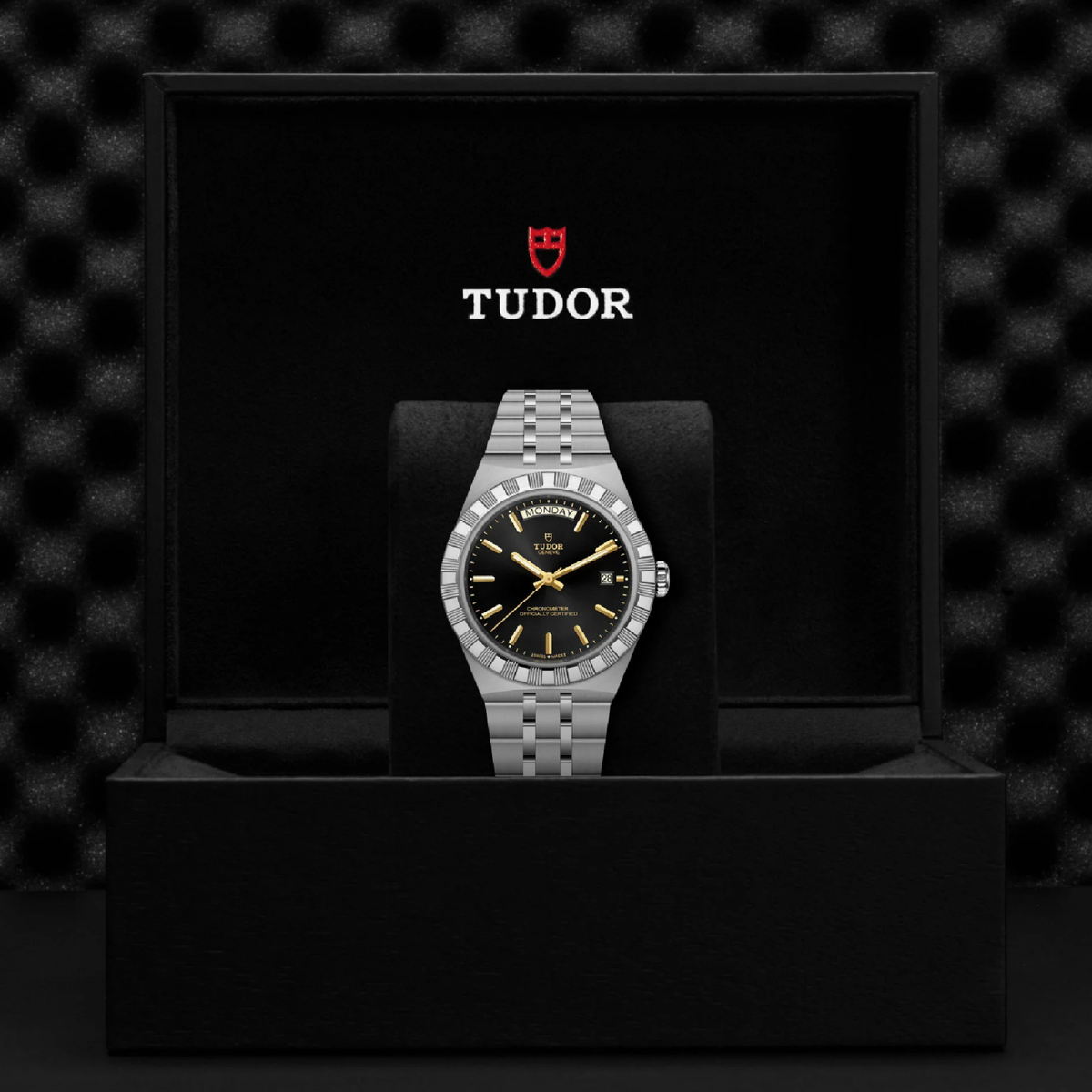 A Tudor watch displayed inside an open black presentation box. The watch has a silver-toned two-tone metal bracelet and case, a black dial with gold hands and markers, and a bezel set with small clear stones.