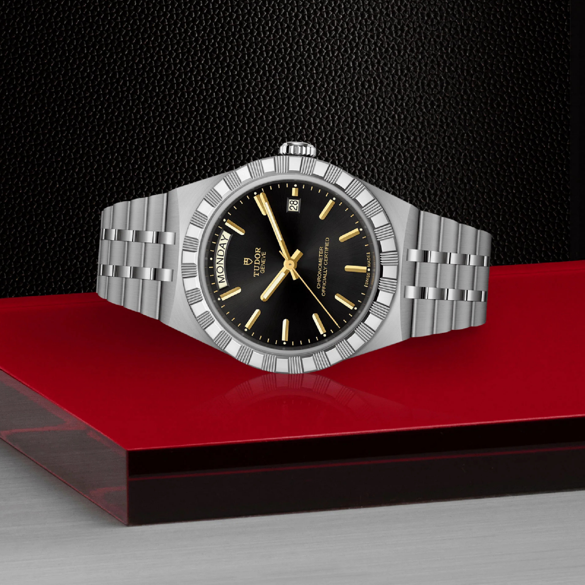 A silver-tone stainless steel wristwatch with a black dial and gold-tone hour markers, displayed on a red platform against a black textured background.