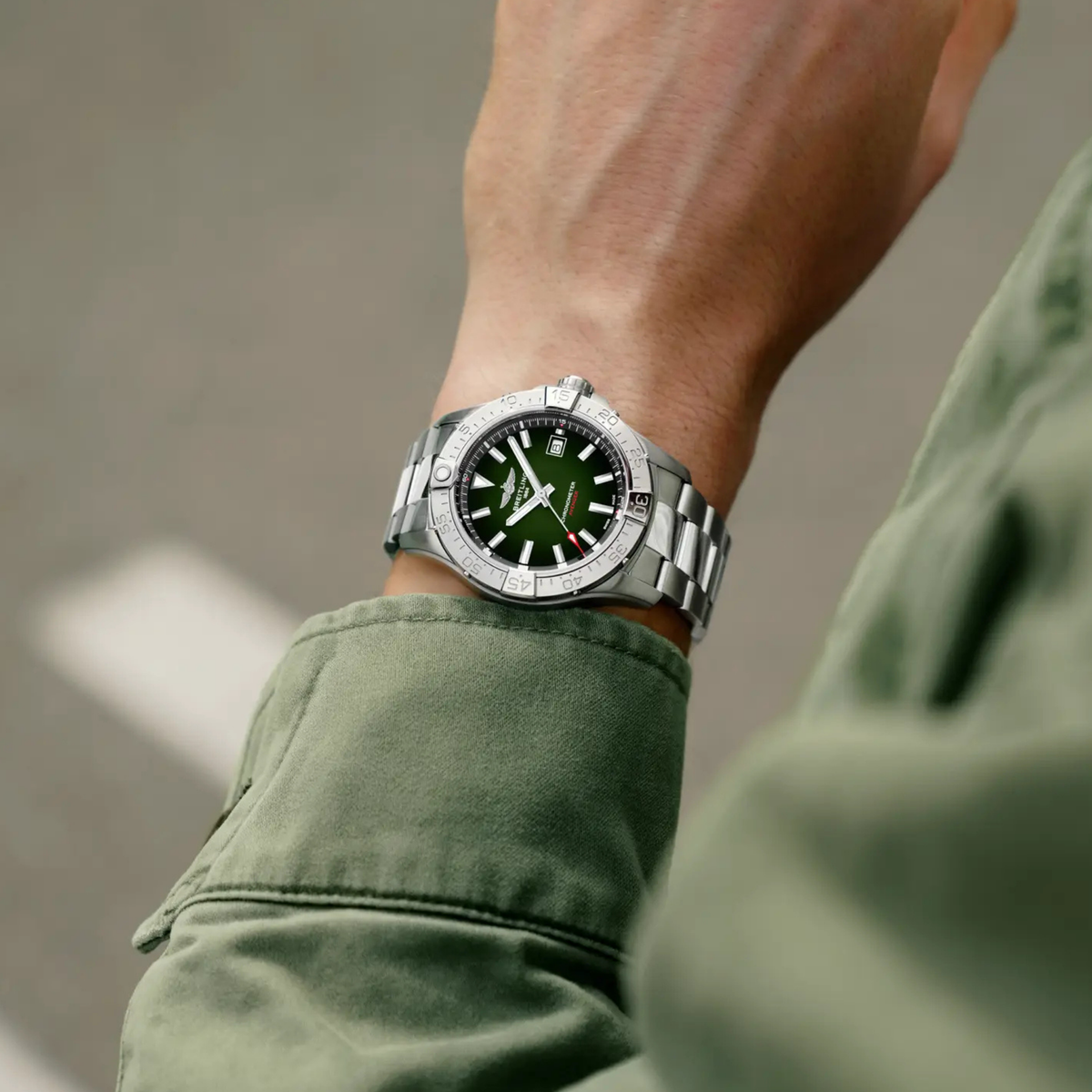 A silver metal wristwatch with a dark green dial and bezel, worn on a person's wrist. The person is wearing an olive green jacket, and the background is a blurred gray pavement.