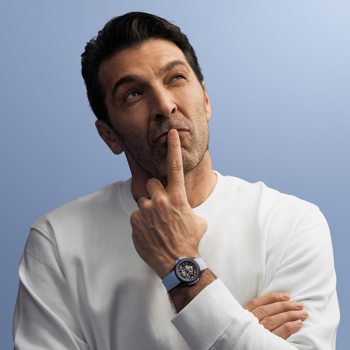 Man wearing a white shirt and blue watch with a thoughtful expression against a light blue background Swiss-made Norqain skeleton watch with a dark textured case, light blue integrated rubber strap and open-worked movement display highlighting exceptional mechanical craftsmanship.