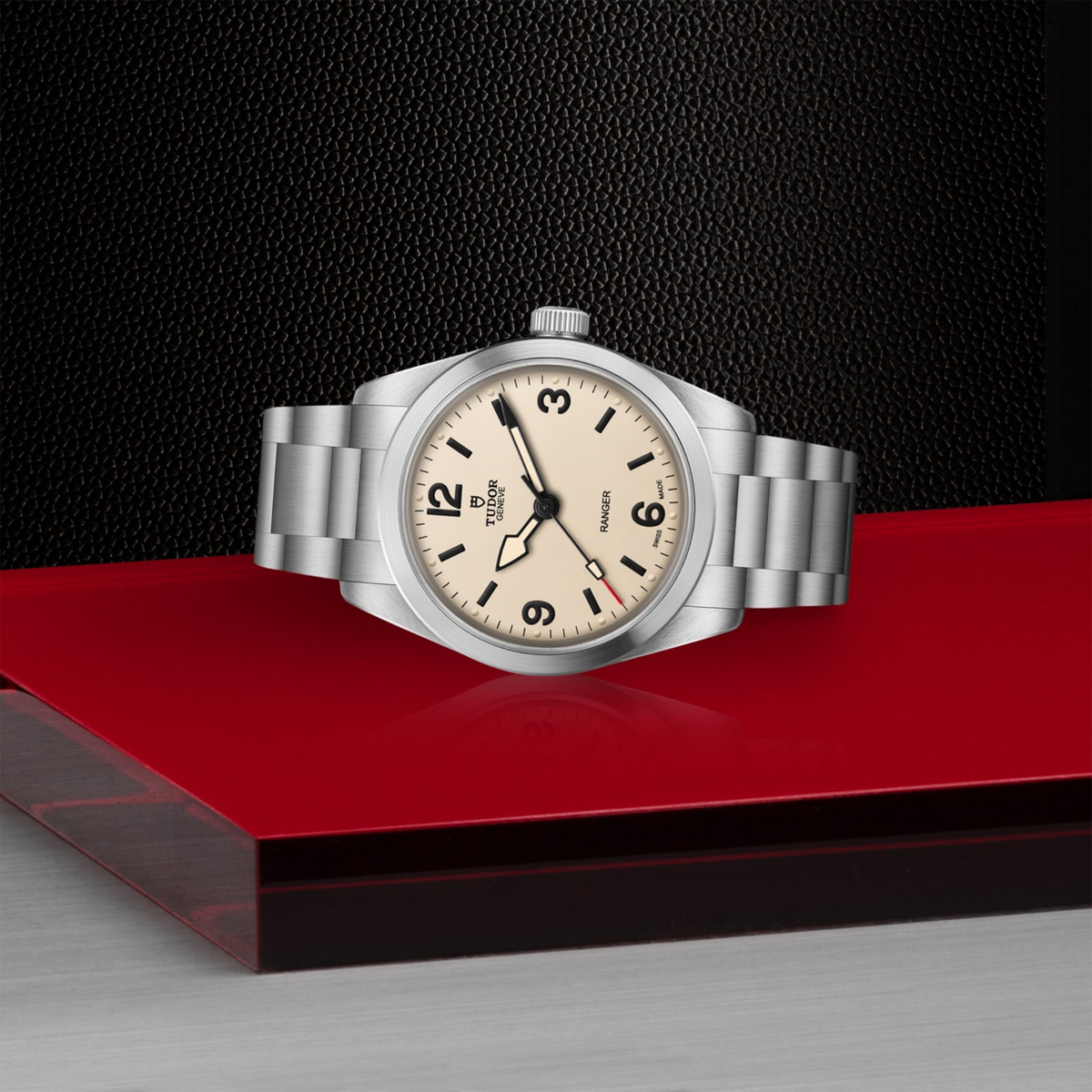 Tudor Ranger watch displayed on a red surface, highlighting its cream dial and stainless steel bracelet.